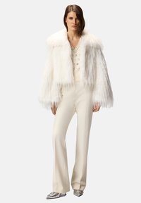 Giacca in pelliccia ecologica bianca con colletto ampio, abbinata a un maglione lavorato a maglia color crema con bottoni e pantaloni svasati color crema. L'outfit è completato da ballerine argento.