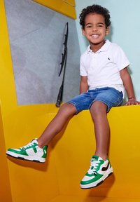 Groene en witte sneakers met zwarte accenten, met een gestructureerde zool en vetersluiting. Draag ze met een spijkerbroekshort en een witte polo.