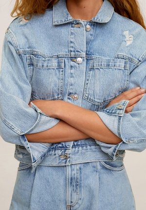 Personne aux bras croisés portant une veste en jean bleu clair délavée et un jean taille haute assorti.