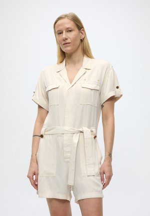 Lyshåret person iført cremefarvet kortærmet romper med bælte, brystlommer og knappedetaljer, stående mod en ensfarvet baggrund.