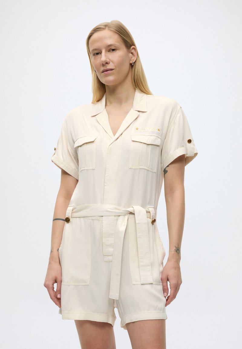 Personne blonde portant une combinaison-short ceinturée à manches courtes de couleur crème avec des poches poitrine et des détails à boutons, debout devant un fond uni.