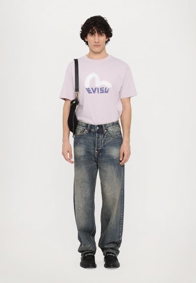 Heled lilla T-särk, millel on "EVISU" logo, kulunud sinised teksad, mustad kingad ja must õlakott, valge tausta ees.