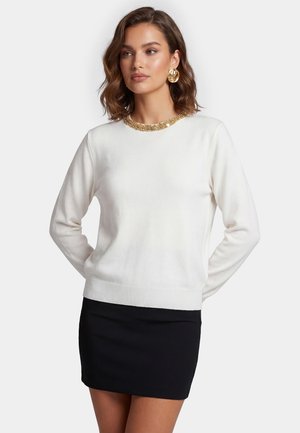 Kvinde iført hvid langærmet sweater med guldkolse med palietter og sort mininederdel, stående med hænderne bag ryggen og kigger til siden.