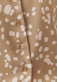 Tan stof blouse met willekeurige witte abstracte vormen. Voorzien van gouden knopen en een gladde textuur.