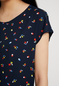 Tmavomodrý top s krátkým rukávem, který má multicolor geometický vzor v červené, bílé, žluté a modré barvě na hladkém materiálu.