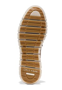 Zool van Cole Haan sneaker, voorzien van een bruine rubberen grip met groeven en richels, en een crèmekleurige rand. Tekst "COLE HAAN GRAND 360" zichtbaar.