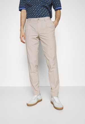 Homme portant un chino beige, des baskets blanches et une chemise à manches courtes à motifs, debout devant un fond blanc uni.