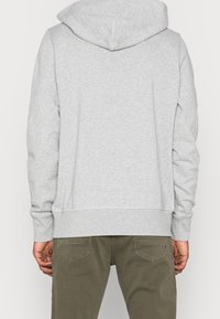 Felpa grigia con una texture morbida, dotata di cappuccio, polsini a coste e orlo dritto. Indossata con pantaloni verde oliva, che mostrano un design pratico.