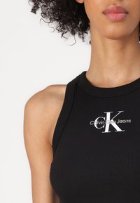 Crna rebrasta majica bez rukava s okruglim izrezom i bijelim logotipom "Calvin Klein Jeans" u sredini. Glatka tkanina s minimalnim detaljima.