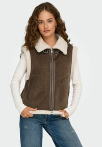Brun kordfløjlsvest med en cremefarvet faux shearling krave, frontlynlås og sidelommer, parret med en hvid langærmet skjorte og blå jeans.