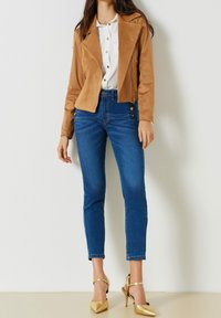 Hnědá faux semišová bunda, zapínání na zip, klopový límec; bílá košile s knoflíky; modré skinny džíny s knoflíkovými detaily; zlaté špičaté jehly.