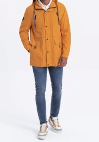 Veste imperméable orange avec capuche, fermeture éclair et boutons-pression à l'avant, deux poches poitrine, et cordons de serrage. Associée à un jean bleu et des baskets multicolores.