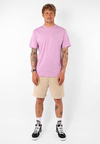 T-shirt rose clair à manches courtes avec un petit logo bleu. Associé à un short beige et des baskets montantes noires avec des lacets blancs.