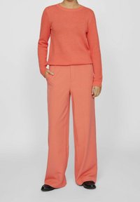 Osoba oblečená v korálovém strukturovaném svetr s dlouhým rukávem a shodnými širokými kalhotami, s černými botami, stojící na jednoduchém pozadí.