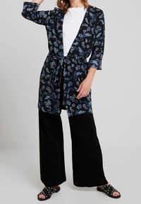 Sort kimono med paisley-mønster og trekvartærmer, bundet i taljen, båret over en hvid t-shirt og parret med brede sorte bukser.