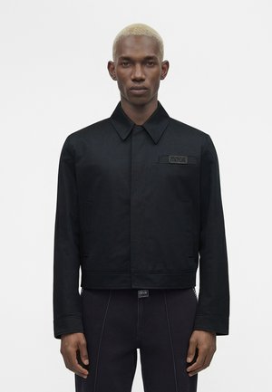 Mees, kellel on lühikesed blondid juuksed, must kraega jakk ja mustad püksid, seisab ühevärvilise valge tausta ees.