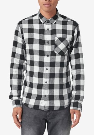 Man die een zwart-wit geruit overhemd met knopen en een voorzak draagt, en donkergrijze spijkerbroek, staand met ontspannen handen.