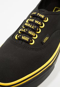 Musta kangaskengät keltaisilla yksityiskohdilla, joissa on ainutlaatuiset nauhat, joissa on tekstiä, ja kumipohja, jossa on erottuva rakenne ja reunus.