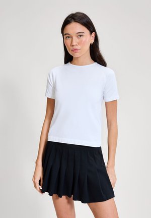 Žena s dlouhými tmavými vlasy, která má na sobě bílou halenku s krátkými rukávy a černou plisovanou sukni, stojící před jednobarevným světlým pozadím.