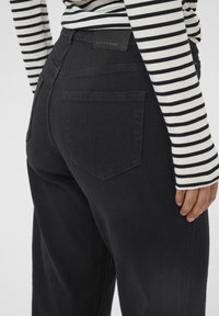 Personne portant un jean en denim noir taille haute avec une étiquette en cuir de marque et une chemise à manches longues rayée noire et blanche.