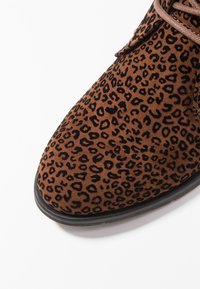 Scarpa in suede marrone con motivo a stampa leopardata nera, forma a punta e lacci marroni, su sfondo bianco.