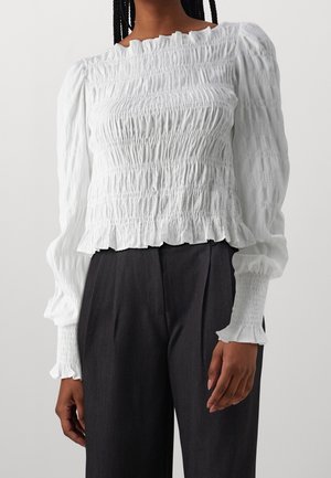 Vrouw draagt een witte blouse met lange mouwen en smockwerk met gerimpelde zoom, in combinatie met zwarte broek met hoge taille, getoond van schouders tot heupen.
