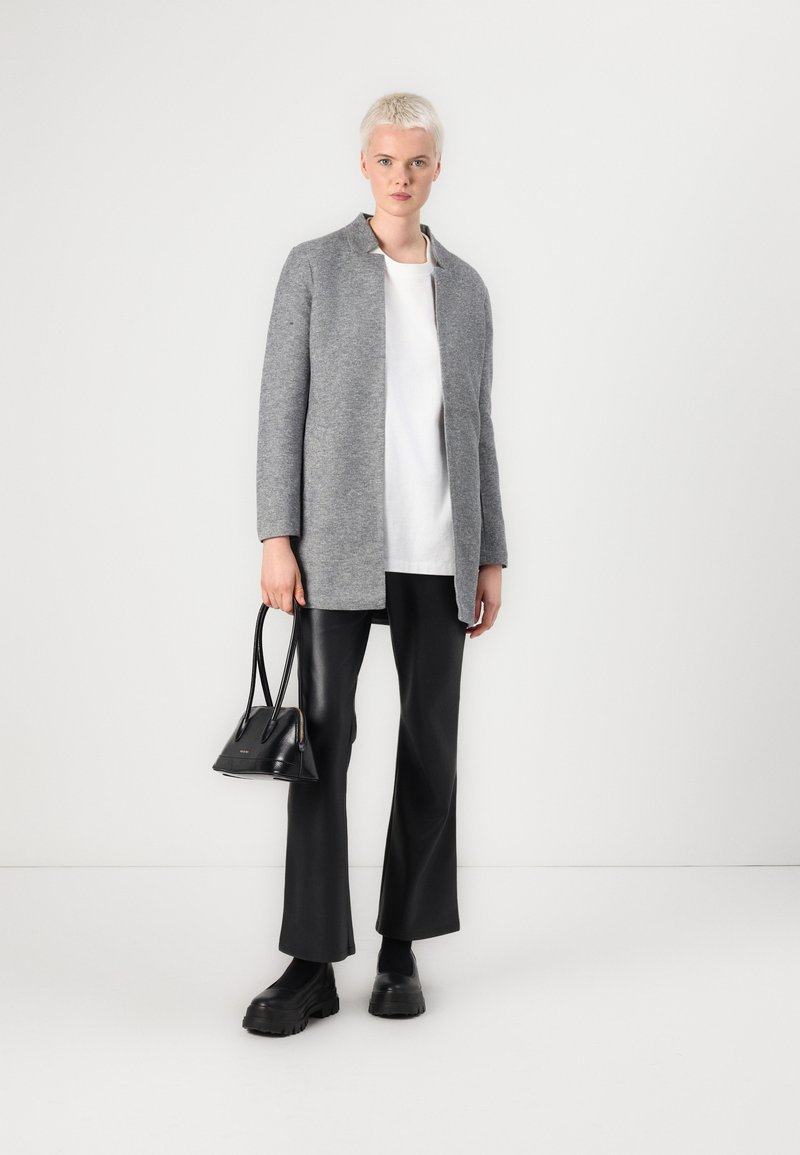 Grau gestrickte Blazerjacke aus Wollmischung mit Kragen, getragen über einem weißen Oberteil, kombiniert mit schwarzen Lederhosen und klobigen schwarzen Schuhen, und hält eine kleine schwarze Tasche.