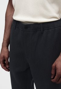 Gros plan d'une personne portant un pantalon noir avec un bouton et des passants de ceinture, associé à une chemise en maille texturée blanche.