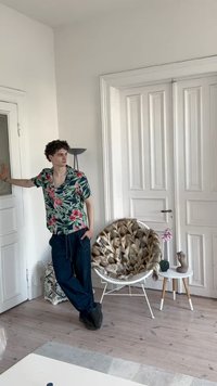 Jaunas vyras gėlėtu marškinėliu ir tamsiomis kelnėmis remiasi į baltas duris kambaryje su kailiniu užklotu krėslu ir mažuoju staliuku su orchidėja.