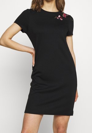 Schwarzes kurzärmliges Kleid aus glattem Stoff, mit rosa Blumenstickerei auf der linken Schulter und figurbetonter Silhouette.