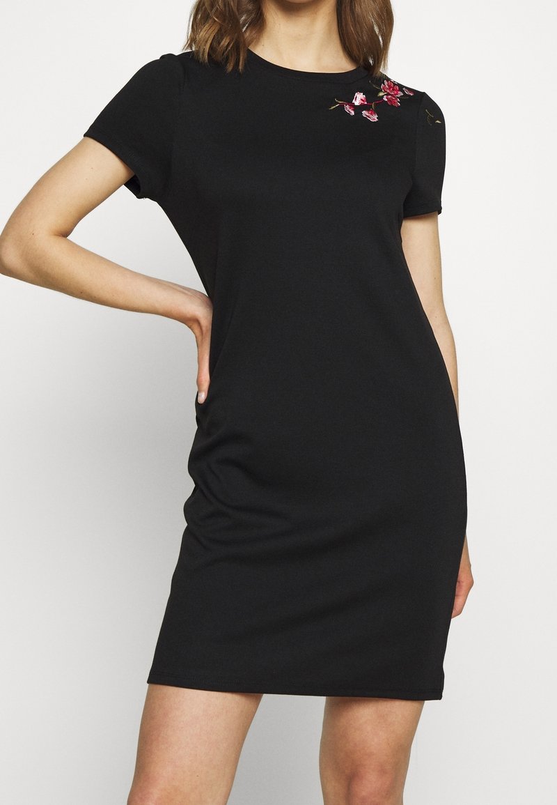 Robe noire à manches courtes en tissu lisse, ornée d'une broderie florale rose sur l'épaule gauche et affichant une silhouette ajustée.