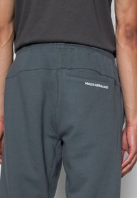 Graue Jogginghose mit elastischem Bund, zwei Gesäßtaschen und weißem "MADS NØRGAARD" Schriftzug auf der rechten Seite. Glatte Textur.