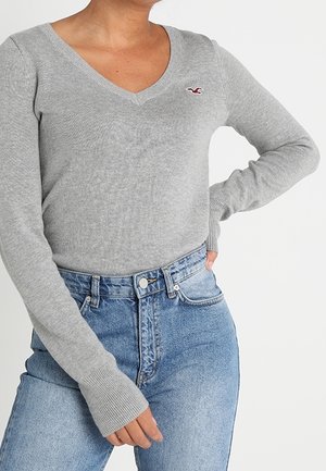Woman wearing a fitted grey V-neck sweater with long sleeves and high-waisted blue jeans, hand resting on hip.