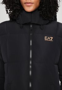 Giubbotto imbottito nero con colletto alto e cappuccio, dotato di zip frontale e logo EA7 in bronzo sottile. Texture liscia e design strutturato.