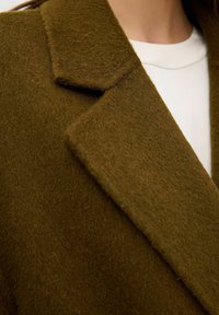 Olivgrön ullblandad kappa med slagkrage, textil med struktur och ett enkelt vitt innerlager som betonar värme och mjukhet.