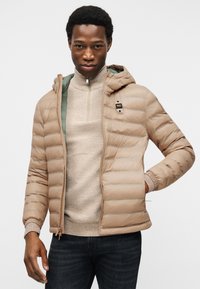 Άνδρας που φοράει μπεζ μπουφάν με κουκούλα και επένδυση πάνω από μπεζ πουλόβερ με μισό φερμουάρ και σκούρο τζιν, στέκεται μπροστά σε ένα απλό φωτεινό φόντο.