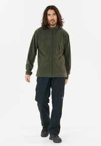 Chaqueta de forro polar en verde oscuro con cuello alto, cremallera frontal y bolsillos laterales; combinada con pantalones negros y zapatos de senderismo.