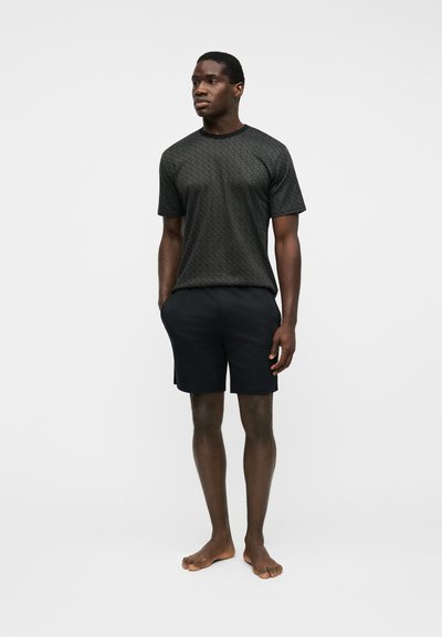 Homme debout pieds nus, faisant face légèrement à gauche, portant une chemise sombre à manches courtes avec des motifs et un short noir arrivant aux genoux sur un fond blanc.