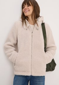 Femme souriante portant une veste à capuche en sherpa crème zippée et un jean bleu, tenant un sac à épaule matelassé vert olive.