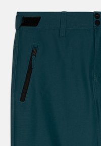 Turkizne tkaninske hlače z dżepom z zadrgo na strani, ki imajo črno zadrgo in velcro okras na pasu.