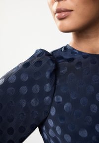 Marineblaue durchsichtige Bluse mit großen, strukturierten Punkten. Verfügt über aufgebauschte Ärmel und einen glatten Ausschnitt, der den subtilen Glanz des Stoffs betont.