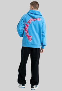 Jasnoniebieska bluza z kapturem z wyraźnym różowym napisem "Combat Heritage" z tyłu, o luźnym kroju i ściągaczach. W połączeniu z czarnymi spodniami.