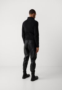 Hombre vistiendo un suéter de canalé negro, pantalones jogger de cuero negro y botas Chelsea negras, de pie de espaldas contra un fondo blanco.