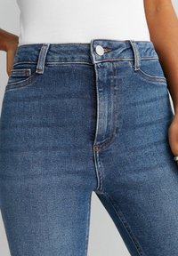 Höga blå denimjeans med ljusa slitageeffekter, kontrasterande sömmar och en slät textur. Har frampocketar och metalldragkedja.