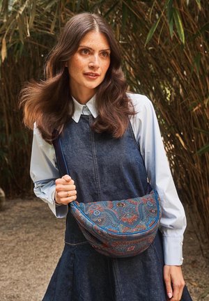 Vrouw met lang bruin haar, gekleed in een lichtblauw shirt en een spijkerjurk, die een blauwe schoudertas met patroon vasthoudt, buiten bij hoge planten.