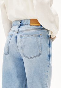 Vaaleansiniset farkut, joissa on korkea vyötärö, näkyvät saumat, kaksi takataskua ja vaaleanruskea nahkainen merkki vyötäröllä.