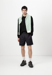 Chemise noire à manches longues, shorts sombres, pull vert et blanc à motifs drapé sur les épaules, chaussures de sport noires et grises, chaussettes blanches.