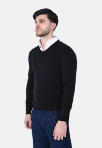 Giovane uomo con capelli scuri e barba, che indossa un maglione nero a V sopra una camicia bianca con colletto e jeans blu, in piedi su uno sfondo bianco.