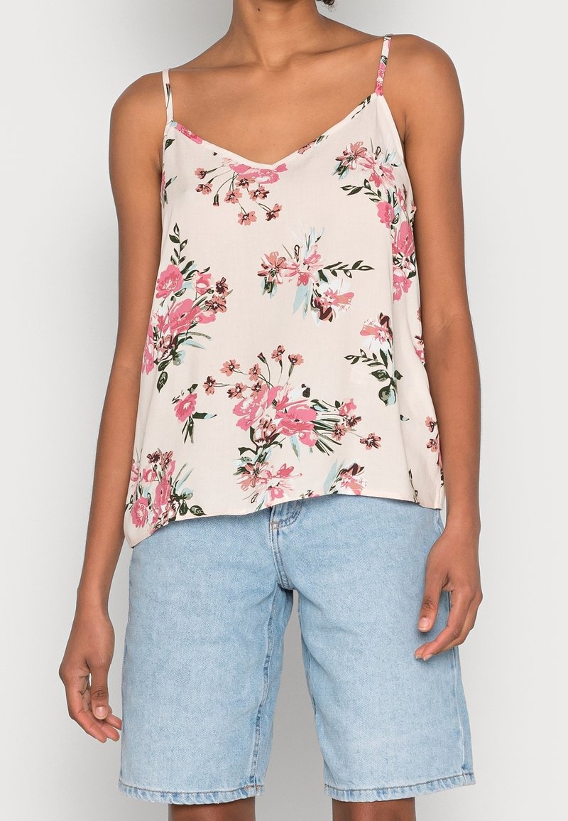 Donna con un top bianco senza maniche con motivo floreale rosa e pantaloni corti in denim blu chiaro fino al ginocchio, in piedi su uno sfondo semplice.