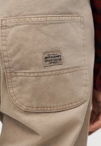 Τσέπη σε μπεζ παντελόνι με ραμμένη ετικέτα που γράφει «Jack & Jones Cotton Dry Goods Div Established 1990» στο ισχίο ενός ατόμου.